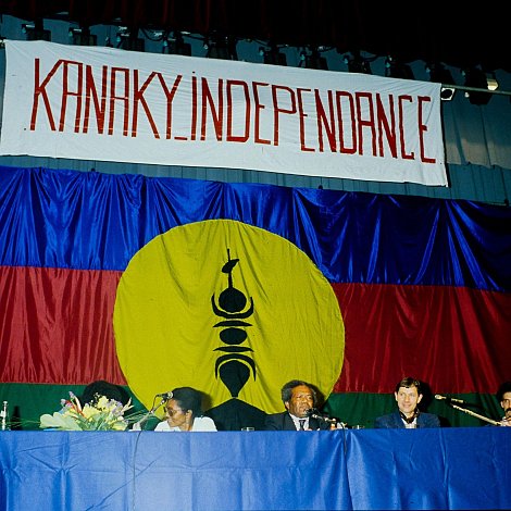 L'image présente une scène de réunion ou de conférence, où des personnes sont assises derrière une table. En arrière-plan, il y a un grand drapeau avec des couleurs vives : un rouge et un bleu, et un cercle jaune au centre. Dans ce cercle, on peut distinguer un symbole noir, précisant ainsi l'identité culturelle. Au-dessus de la scène, une banderole arbore le mot "KANAKY INDEPENDANCE" écrit en lettres rouges, exprimant un message fort lié à l'indépendance. Les participants semblent engagés et sont en discussion, créant une atmosphère de détermination et de solidarité.