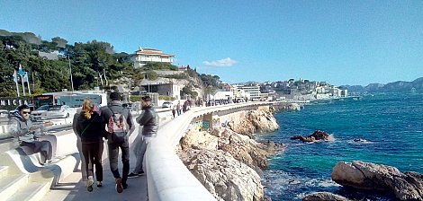 Imaginez-vous sur une promenade longeant la mer. Sur votre droite, vous pouvez entendre le bruit des vagues qui s'écrasent doucement contre les rochers, tandis que l'odeur salée de l'océan flotte dans l'air. Le vent léger caresse votre visage et apporte une sensation de fraîcheur. À votre gauche, des bâtiments se dressent, certains sont des maisons et d'autres des commerces. Vous pouvez sentir l'animation de la ville avec des groupes de personnes qui marchent, certains discutant joyeusement. La promenade est large et bien aménagée, invitant à la flânerie. Au loin, le paysage est ponctué d'arbres et de collines, avec quelques nuages blancs dégradant le bleu du ciel. La lumière du soleil illumine tout le paysage, créant une ambiance chaleureuse. L'ensemble évoque une belle journée au bord de la mer, propice à la détente et au plaisir des sens.